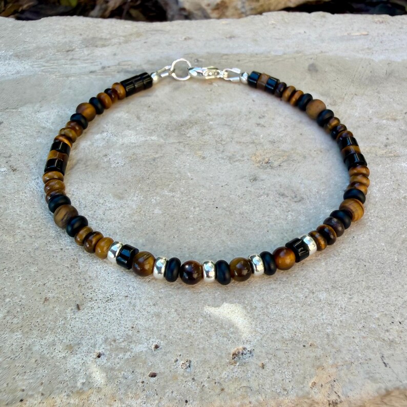 K&ouml;nnte beinhalten: Ein Perlenarmband mit abwechselnden runden Tigeraugen-Perlen, mattschwarzen zylindrischen Perlen und kleinen silberfarbenen Metall-Abstandshaltern. Das Armband wird mit einem silberfarbenen Karabinerverschluss und einem Biegering geschlossen.