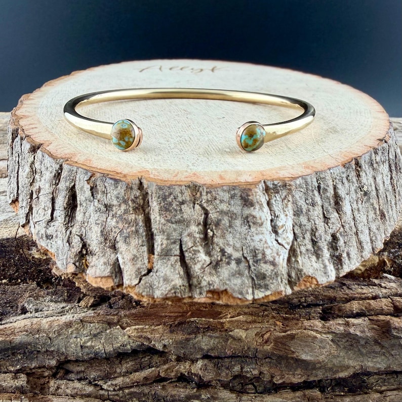 May include: A gold-colored cuff bracelet with two turquoise-colored stones. The bracelet is displayed on a rustic wooden surface, showcasing its elegant design and natural elements. The stones are round and set in gold bezels.