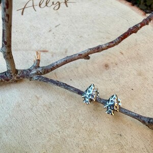 May include: A pair of silver-colored arrowhead stud earrings. The earrings are detailed with a textured surface and are displayed on a small tree branch against a light wood background. The word "Ally" is written in cursive in the upper left corner.