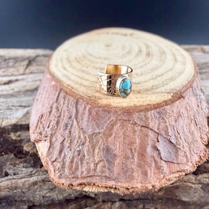 May include: A gold-colored ring with a turquoise stone. The ring is set on a wooden surface. The ring has an engraved design on the side and a bright blue and brown oval-shaped stone. The ring is a piece of jewelry.