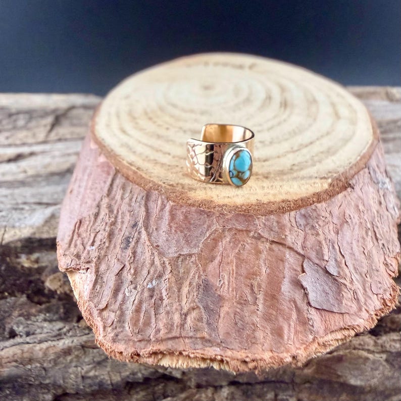 May include: A gold-colored ring with a turquoise stone. The ring is set on a wooden surface. The ring has an engraved design on the side and a bright blue and brown oval-shaped stone. The ring is a piece of jewelry.