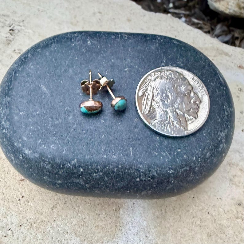 May include: A pair of gold-colored stud earrings with turquoise-colored stones. The earrings are displayed on a dark gray stone next to a silver coin. The earrings have a post and backing.