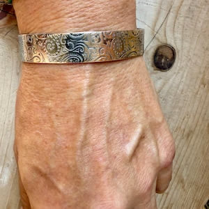 May include: A silver-toned metal cuff bracelet featuring an intricate, repeating embossed pattern of swirling lines, spirals, and small star shapes across the entire outer surface. The bracelet has a flat, wide band design with a polished finish.