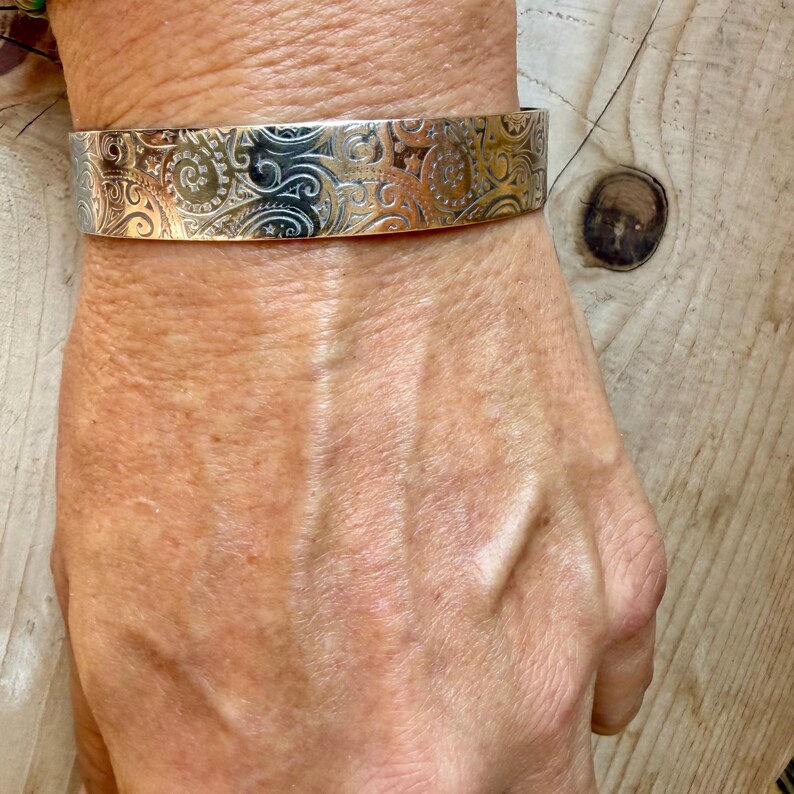 May include: A silver-toned metal cuff bracelet featuring an intricate, repeating embossed pattern of swirling lines, spirals, and small star shapes across the entire outer surface. The bracelet has a flat, wide band design with a polished finish.