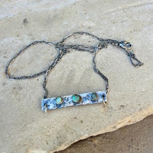 May include: A silver necklace featuring a hammered bar pendant with three turquoise stones. The pendant is connected to a chain necklace with a lobster clasp. The necklace is displayed on a light gray surface.