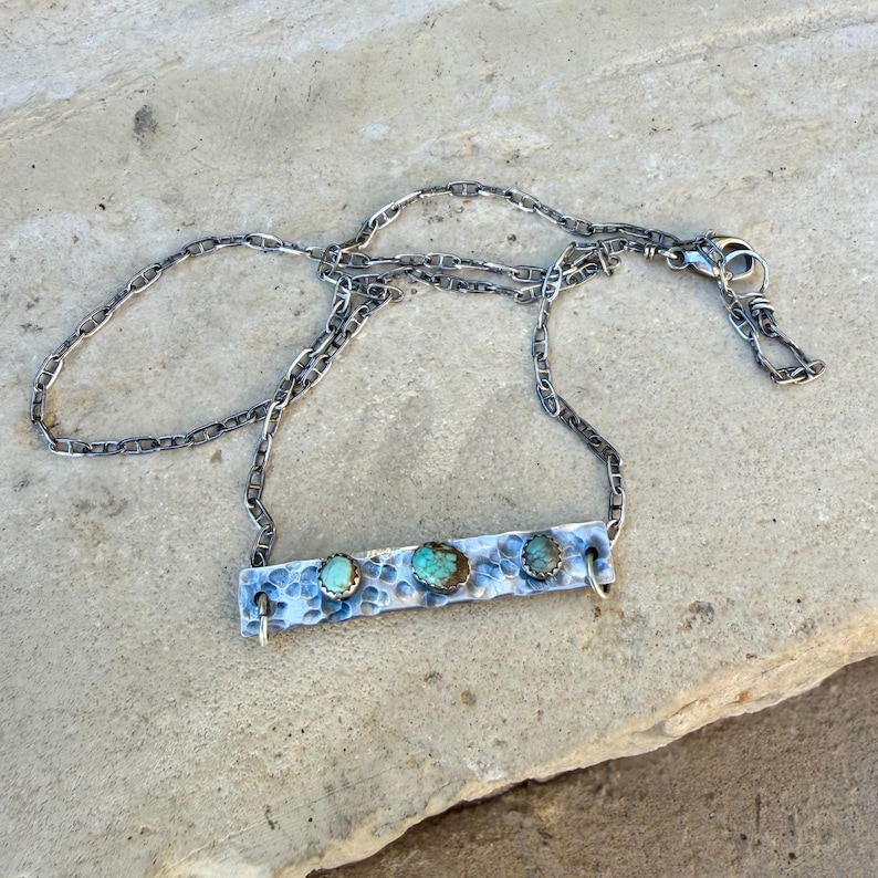 May include: A silver necklace featuring a hammered bar pendant with three turquoise stones. The pendant is connected to a chain necklace with a lobster clasp. The necklace is displayed on a light gray surface.