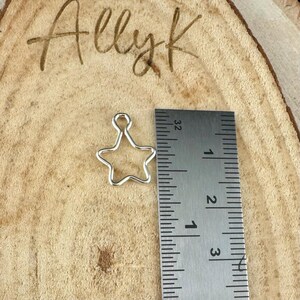 May include: A small, silver-colored star-shaped charm with a loop at the top. The charm is approximately 0.75 inches in height. The charm is displayed on a wooden surface next to a ruler.