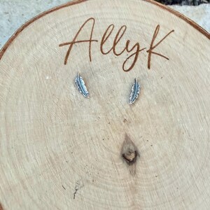 May include: Two small, silver-colored feather earrings displayed on a light brown wooden surface. The earrings are detailed with feather-like textures. The word "Allyk" is written in cursive script on the wood.