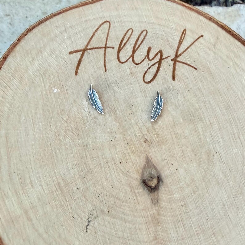 May include: Two small, silver-colored feather earrings displayed on a light brown wooden surface. The earrings are detailed with feather-like textures. The word "Allyk" is written in cursive script on the wood.