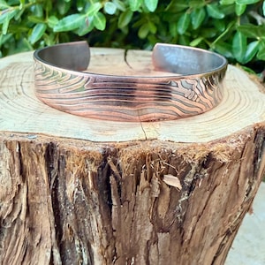 May include: Copper cuff bracelet with a textured, engraved design. The bracelet features a wide band with a detailed pattern of lines and shapes. The metal has a warm, reddish-brown hue, with a contrasting silver-colored interior.