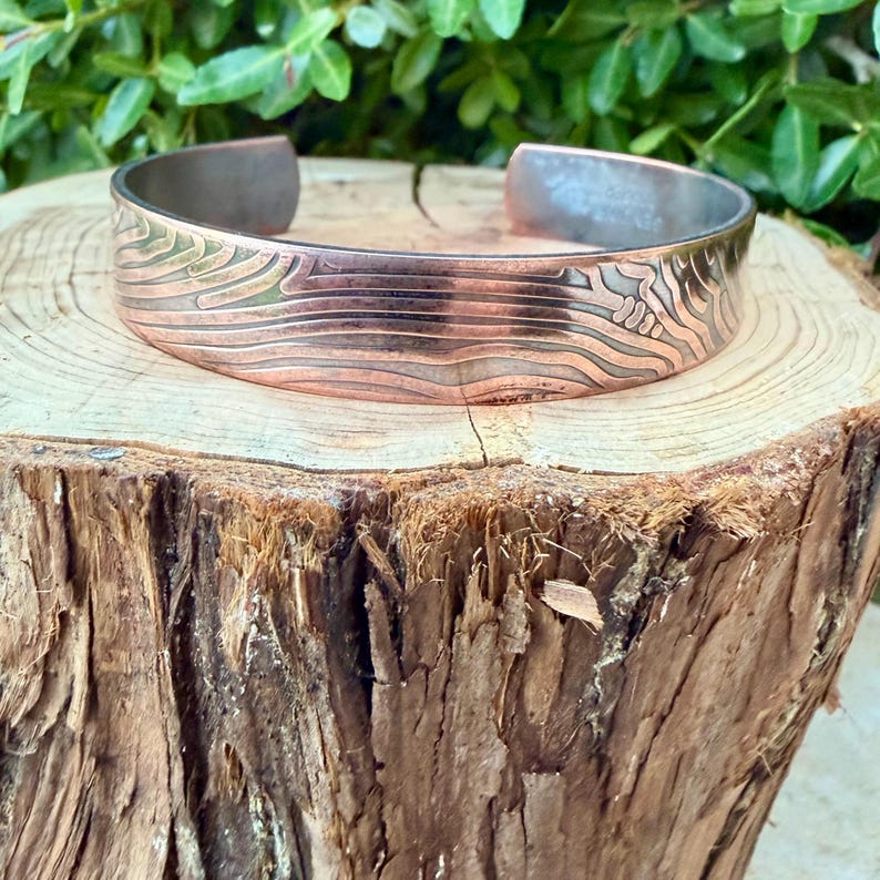 May include: Copper cuff bracelet with a textured, engraved design. The bracelet features a wide band with a detailed pattern of lines and shapes. The metal has a warm, reddish-brown hue, with a contrasting silver-colored interior.