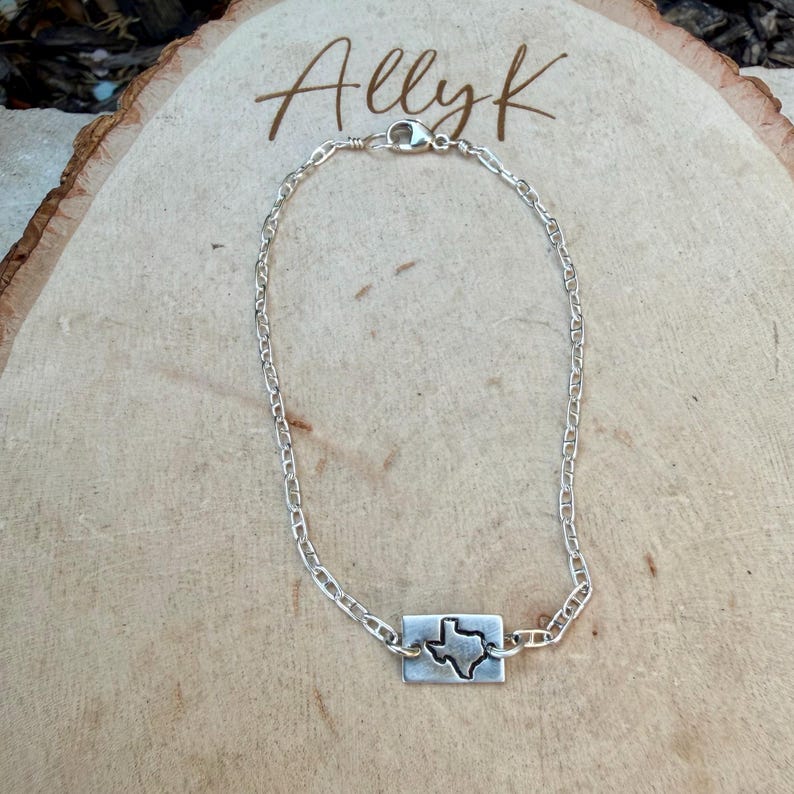 May include: A silver chain bracelet with a rectangular charm featuring a cutout of the state of Texas. The bracelet is displayed on a wooden surface, with the name "Allyk" written in cursive script.