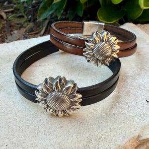 May include: Two leather bracelets, one brown and one black, each featuring a silver sunflower clasp. The bracelets have a double-wrap design and a magnetic closure. The sunflower clasp is detailed and adds a decorative element.