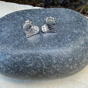 May include: A pair of silver heart-shaped stud earrings featuring an intricate engraved pattern on the surface. Each earring has a polished metallic finish with detailed scrollwork and a small scalloped edge design along the top border.