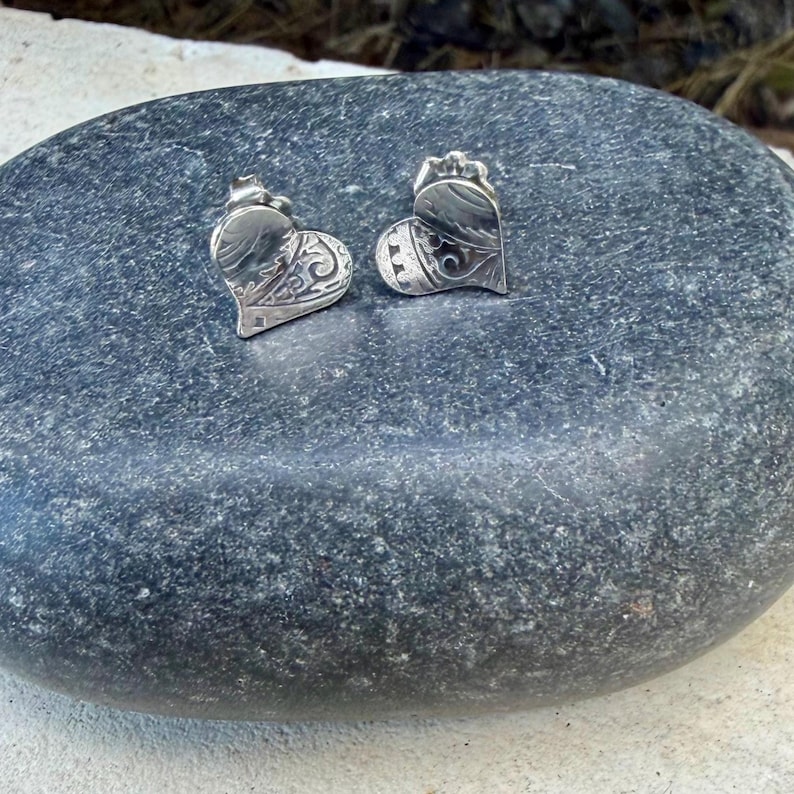May include: A pair of silver heart-shaped stud earrings featuring an intricate engraved pattern on the surface. Each earring has a polished metallic finish with detailed scrollwork and a small scalloped edge design along the top border.
