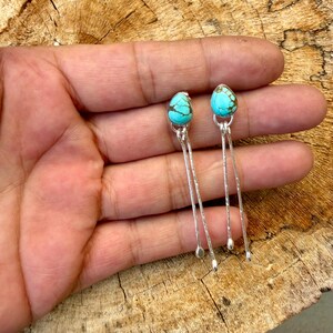 May include: A pair of dangle earrings featuring teardrop-shaped turquoise stones set in silver bezels. Each earring has a long, thin silver wire extending downward from the stone, ending in a small, rounded silver bead at the base of the wire.