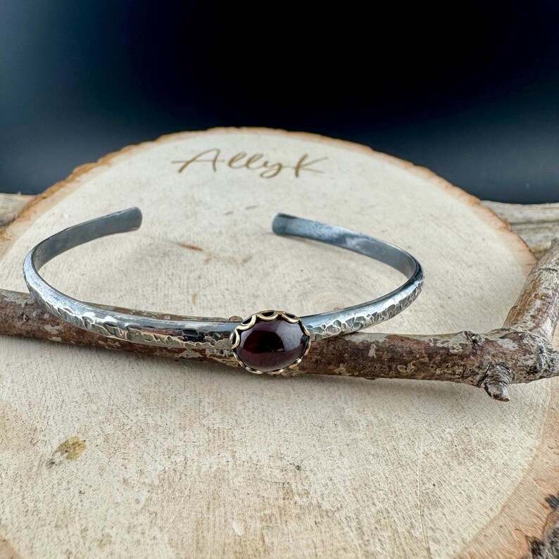 Puede incluir: Una pulsera de plata con textura martillada y una piedra preciosa de color rojo oscuro engastada en un bisel dorado. La pulsera es abierta y descansa sobre una superficie de madera.