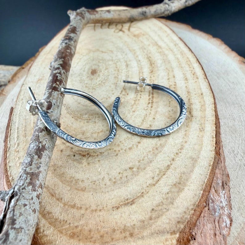 May include: A pair of silver hoop earrings with a detailed, etched pattern. The earrings are displayed on a wooden surface with a natural, rustic aesthetic. The earrings have a post and are a classic jewelry item.