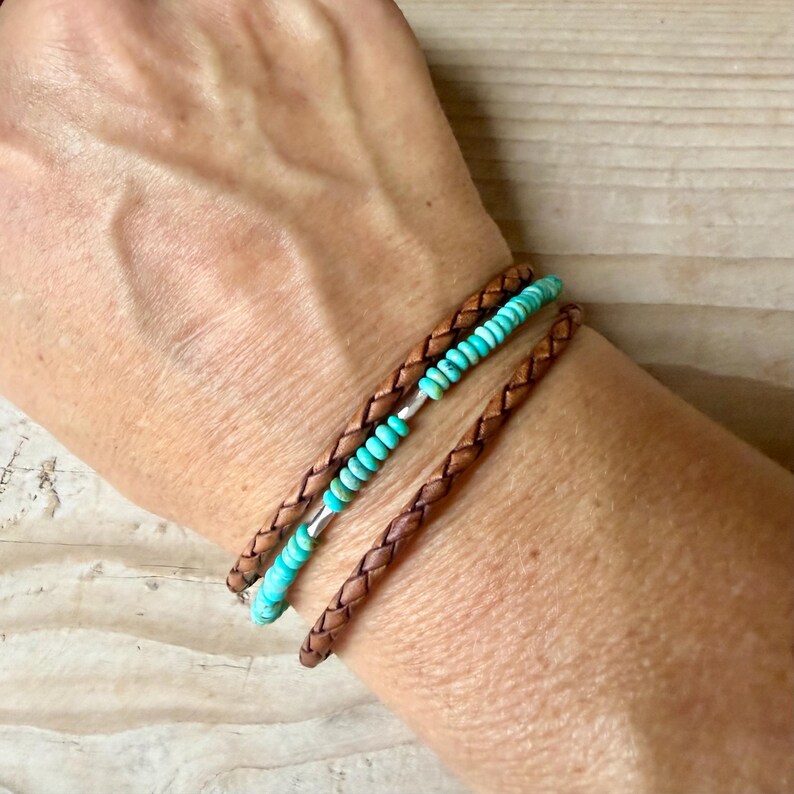 May include: A multi-strand bracelet featuring two braided brown leather cords and a central strand of small, round, turquoise-colored beads. The design incorporates metallic silver-toned accents interspersed among the beads, creating a layered look.