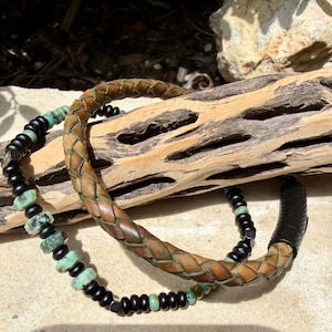 May include: Two bracelets. One is a braided brown leather bracelet. The other is a beaded bracelet with black and turquoise beads. The bracelets are displayed on a piece of driftwood.