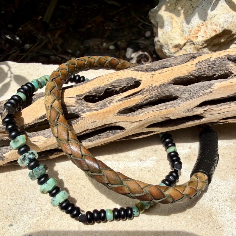 May include: Two bracelets. One is a braided brown leather bracelet. The other is a beaded bracelet with black and turquoise beads. The bracelets are displayed on a piece of driftwood.