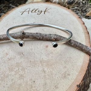 Könnte beinhalten: Ein silberfarbenes, offenes Armband mit einem kleinen schwarzen Stein an jedem Ende. Das Armband wird auf einer Holzoberfläche präsentiert. Der Text "Allyk" ist in das Holz graviert.