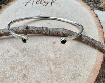 Op de afbeelding: Een zilverkleurige open manchetarmband met een kleine zwarte steen aan elk uiteinde. De armband wordt gepresenteerd op een houten oppervlak. De tekst "Allyk" is in het hout gegraveerd.