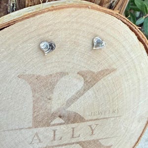 May include: A pair of small, silver-toned heart-shaped stud earrings with a textured, metallic finish. The earrings are displayed on a circular wooden slice featuring the engraved text K ALLY JEWELRY across the center surface.