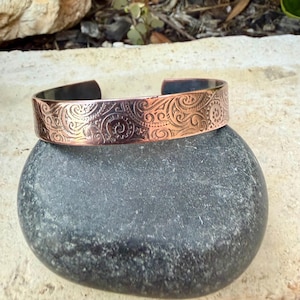 Puede incluir: Brazalete de pu&ntilde;o de cobre con un intrincado dise&ntilde;o de remolinos. El brazalete abierto se muestra sobre una piedra gris lisa. El metal tiene un c&aacute;lido tono marr&oacute;n rojizo, con acentos m&aacute;s oscuros en las zonas hundidas del dise&ntilde;o. El brazalete mide unos 2.5 cm de ancho.