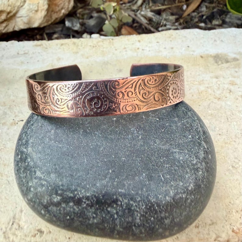 Puede incluir: Brazalete de pu&ntilde;o de cobre con un intrincado dise&ntilde;o de remolinos. El brazalete abierto se muestra sobre una piedra gris lisa. El metal tiene un c&aacute;lido tono marr&oacute;n rojizo, con acentos m&aacute;s oscuros en las zonas hundidas del dise&ntilde;o. El brazalete mide unos 2.5 cm de ancho.