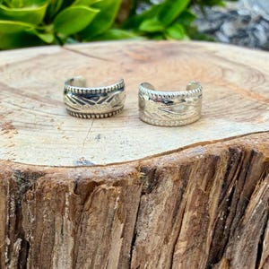 May include: Two silver-toned metal rings with an open-back design. Each ring features a textured surface with a repeating geometric pattern and a notched, beaded border along the top and bottom edges of the band.