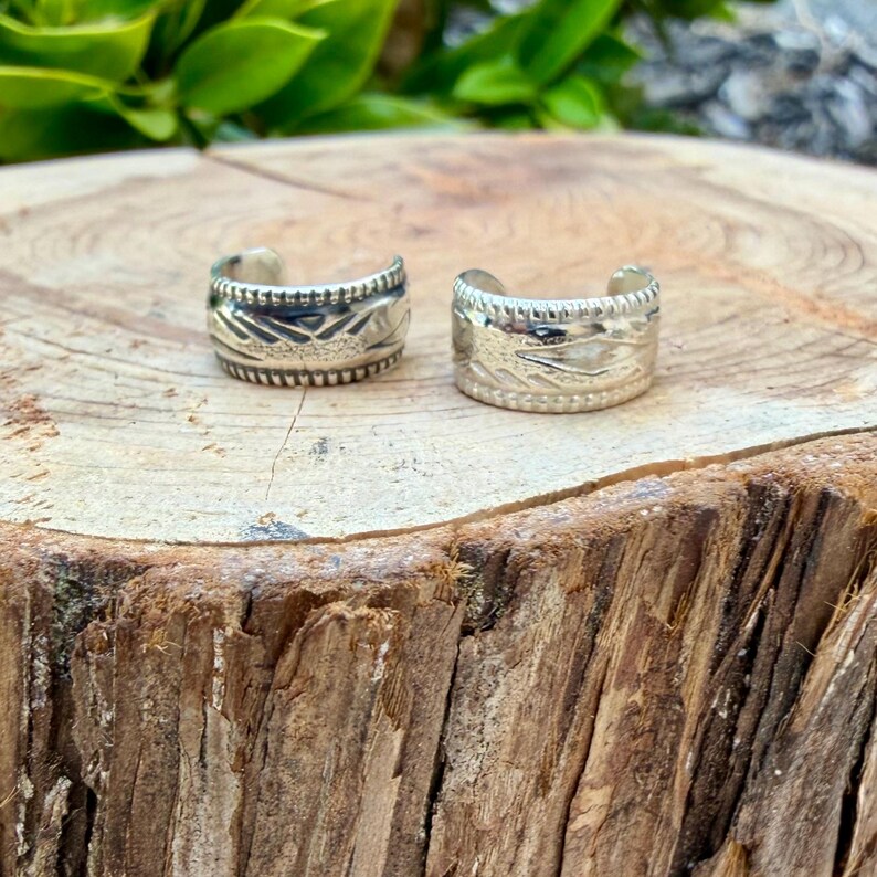 May include: Two silver-toned metal rings with an open-back design. Each ring features a textured surface with a repeating geometric pattern and a notched, beaded border along the top and bottom edges of the band.