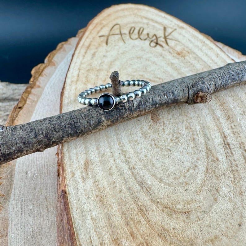 K&ouml;nnte beinhalten: Ein silberner Perlenring mit einem gro&szlig;en, runden, schwarzen Stein in der Mitte. Der Ring wird auf einem kleinen, verwitterten Ast vor einem h&ouml;lzernen Hintergrund pr&auml;sentiert. Der Name "Ally K" ist handschriftlich auf das Holz geschrieben.
