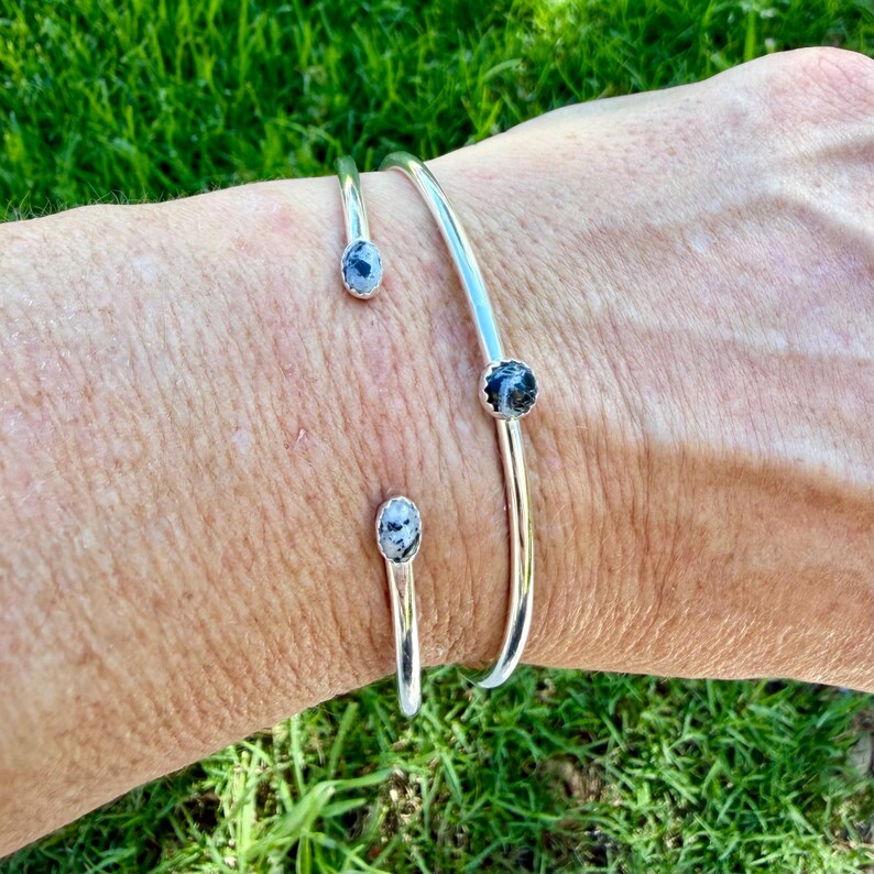 May include: A set of three open-ended silver bracelets. Each bracelet features a small, round, speckled stone in shades of gray and black. The bracelets are displayed on a person's arm against a backdrop of green grass.