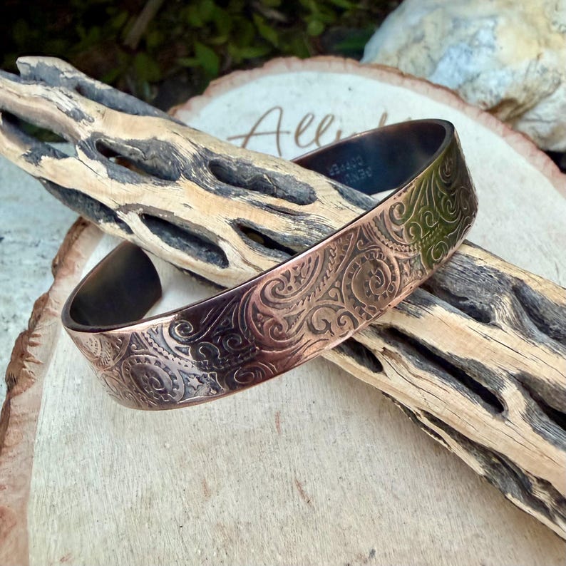 Puede incluir: Una pulsera de cobre con un intrincado patr&oacute;n floral en relieve. La pulsera tiene un acabado oscuro y oxidado en el interior y los bordes, y un tono cobre m&aacute;s claro en el exterior. El dise&ntilde;o presenta l&iacute;neas onduladas y motivos circulares.