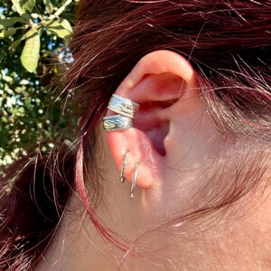 May include: Two silver-toned ear cuffs with a textured, engraved pattern worn on the upper cartilage, paired with two thin, smooth silver-toned hoop earrings positioned in the lower earlobe piercings, creating a layered metal look.