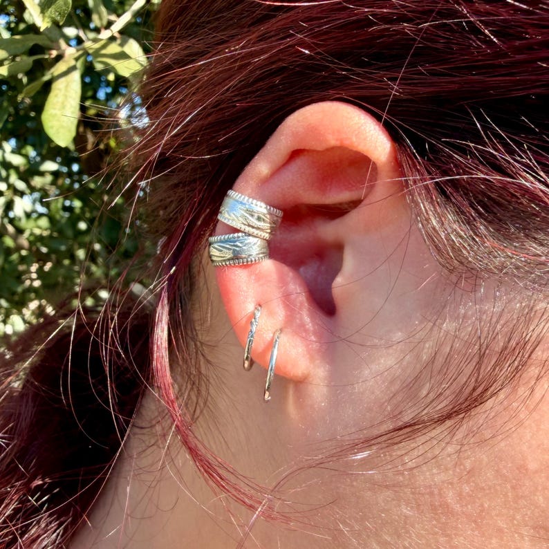 May include: Two silver-toned ear cuffs with a textured, engraved pattern worn on the upper cartilage, paired with two thin, smooth silver-toned hoop earrings positioned in the lower earlobe piercings, creating a layered metal look.