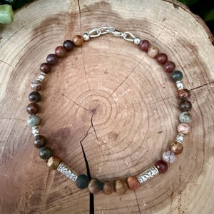 May include: A beaded bracelet featuring round earth-toned stones in shades of brown, tan, and grey, interspersed with small silver-colored metal spacer beads and two larger cylindrical silver-colored metal beads with engraved patterns.