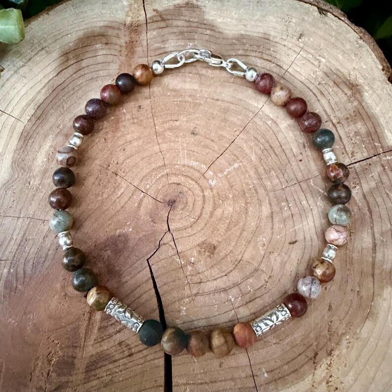 May include: A beaded bracelet featuring round earth-toned stones in shades of brown, tan, and grey, interspersed with small silver-colored metal spacer beads and two larger cylindrical silver-colored metal beads with engraved patterns.
