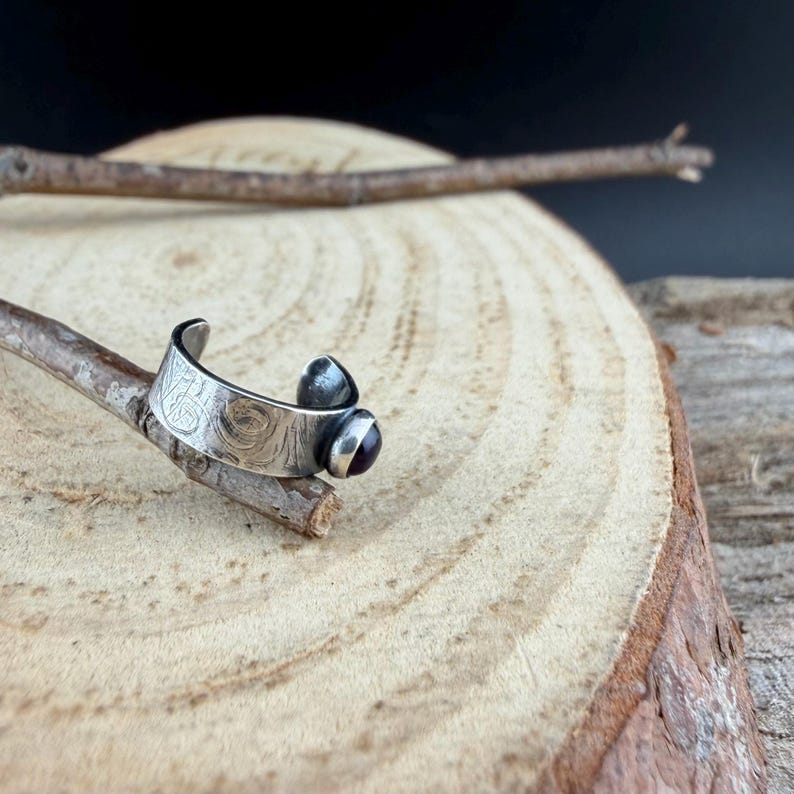 May include: A silver-toned ring with a textured band and a dark purple gemstone. The ring has an open design and is displayed on a small branch against a wooden background. The ring's design features a small, rounded gemstone.