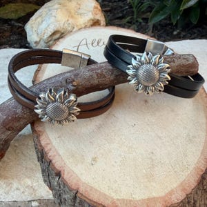 May include: Two leather bracelets, one brown and one black, each featuring a silver sunflower charm. The bracelets have a magnetic clasp. The bracelets are displayed on a wooden surface.