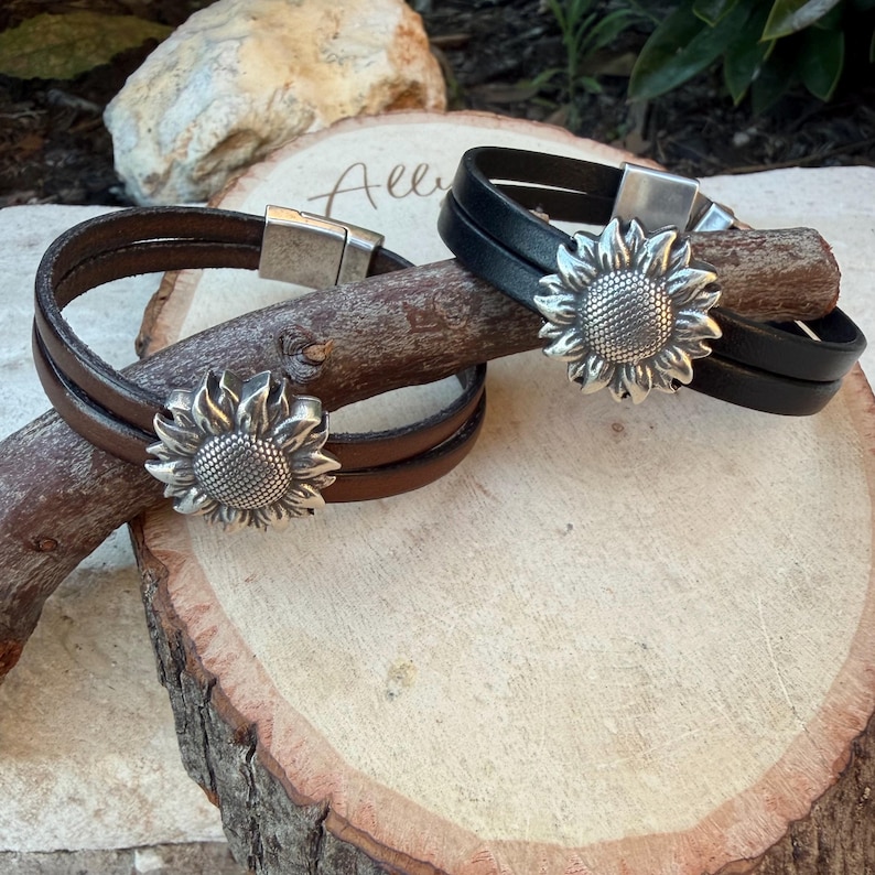 May include: Two leather bracelets, one brown and one black, each featuring a silver sunflower charm. The bracelets have a magnetic clasp. The bracelets are displayed on a wooden surface.