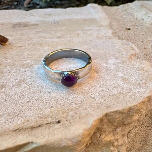 May include: A silver ring with a hammered texture and a round, deep purple gemstone. The ring is set on a light-coloured stone surface, showcasing the contrast between the metal and the jewel.