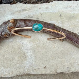 May include: A gold-colored cuff bracelet with a turquoise stone set in the center. The open-ended bracelet is resting on a piece of wood against a light-colored stone surface. The turquoise stone is oval-shaped.