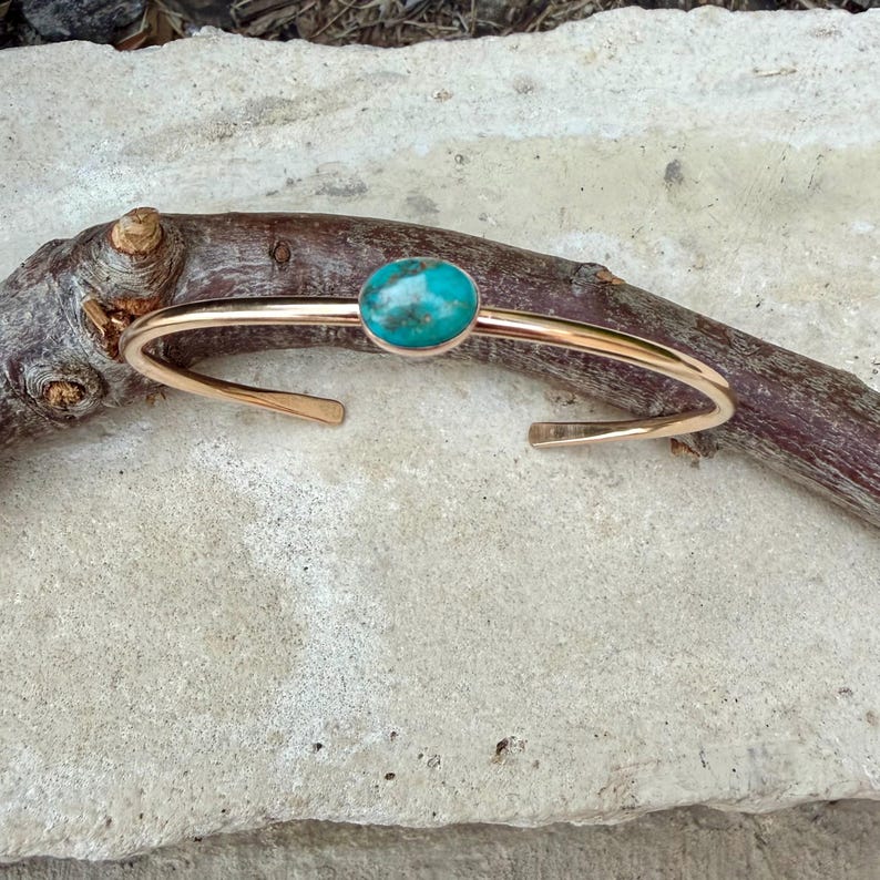 May include: A gold-colored cuff bracelet with a turquoise stone set in the center. The open-ended bracelet is resting on a piece of wood against a light-colored stone surface. The turquoise stone is oval-shaped.