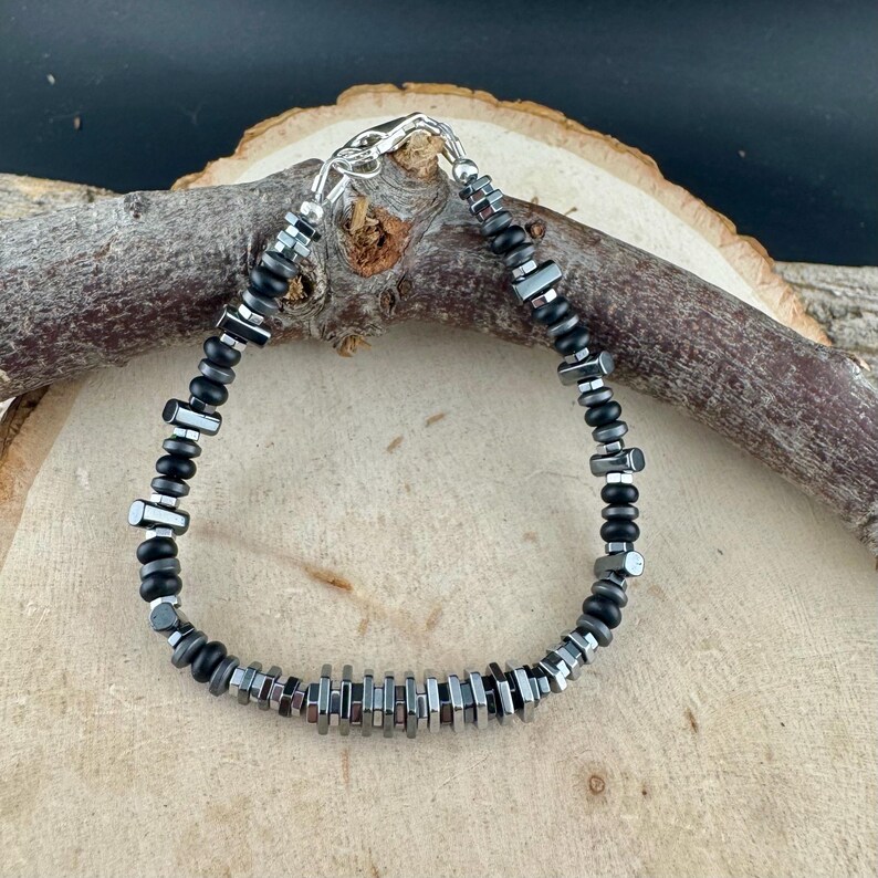 May include: A beaded bracelet featuring a mix of black and silver-toned beads. The bracelet is composed of small, round black beads interspersed with rectangular and cylindrical silver-toned beads. It has a silver clasp.