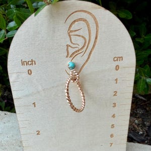 May include: A single earring with a turquoise bead and a copper-colored, twisted oval design. The earring is displayed on a wooden board with inch and centimeter measurements. The earring is approximately 1 inch (2.5 cm) in length.
