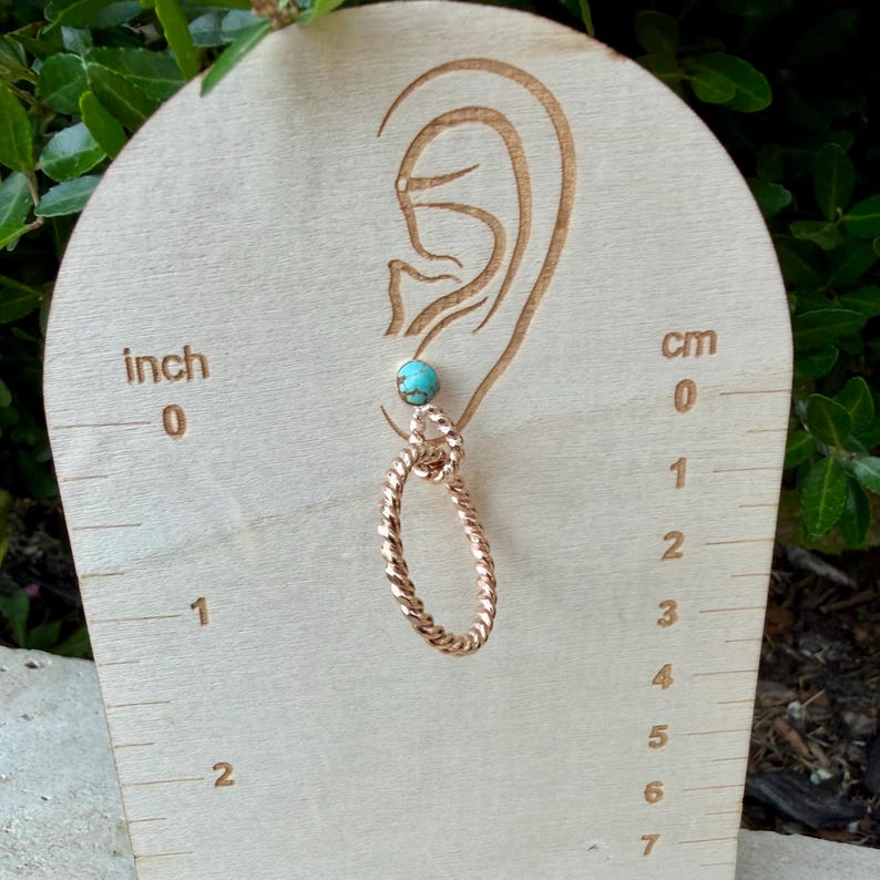 May include: A single earring with a turquoise bead and a copper-colored, twisted oval design. The earring is displayed on a wooden board with inch and centimeter measurements. The earring is approximately 1 inch (2.5 cm) in length.