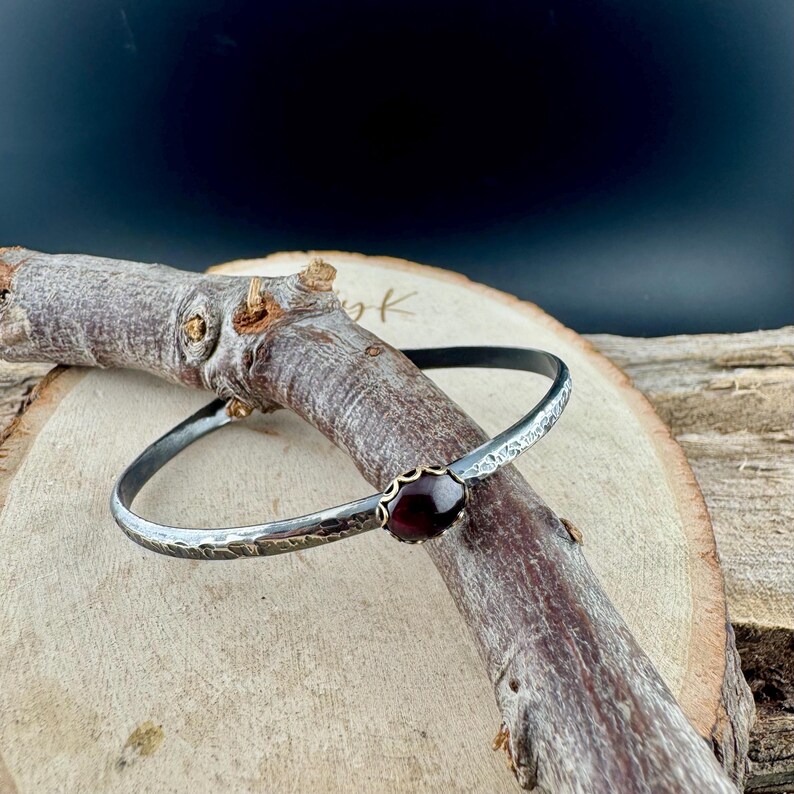 Puede incluir: Un brazalete de plata con textura martillada y una piedra preciosa de color rojo oscuro engastada en un bisel dorado. El brazalete se exhibe sobre una rama de madera sobre un fondo oscuro. La piedra preciosa es ovalada.