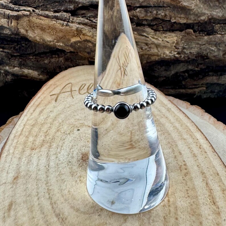 K&ouml;nnte beinhalten: Ein silberner Ring mit einem Perlenband und einem schwarzen, kreisf&ouml;rmigen Stein in der Mitte. Der Ring wird auf einem transparenten, konischen St&auml;nder vor einem Holzgrund pr&auml;sentiert. Das Design des Rings ist schlicht und elegant.